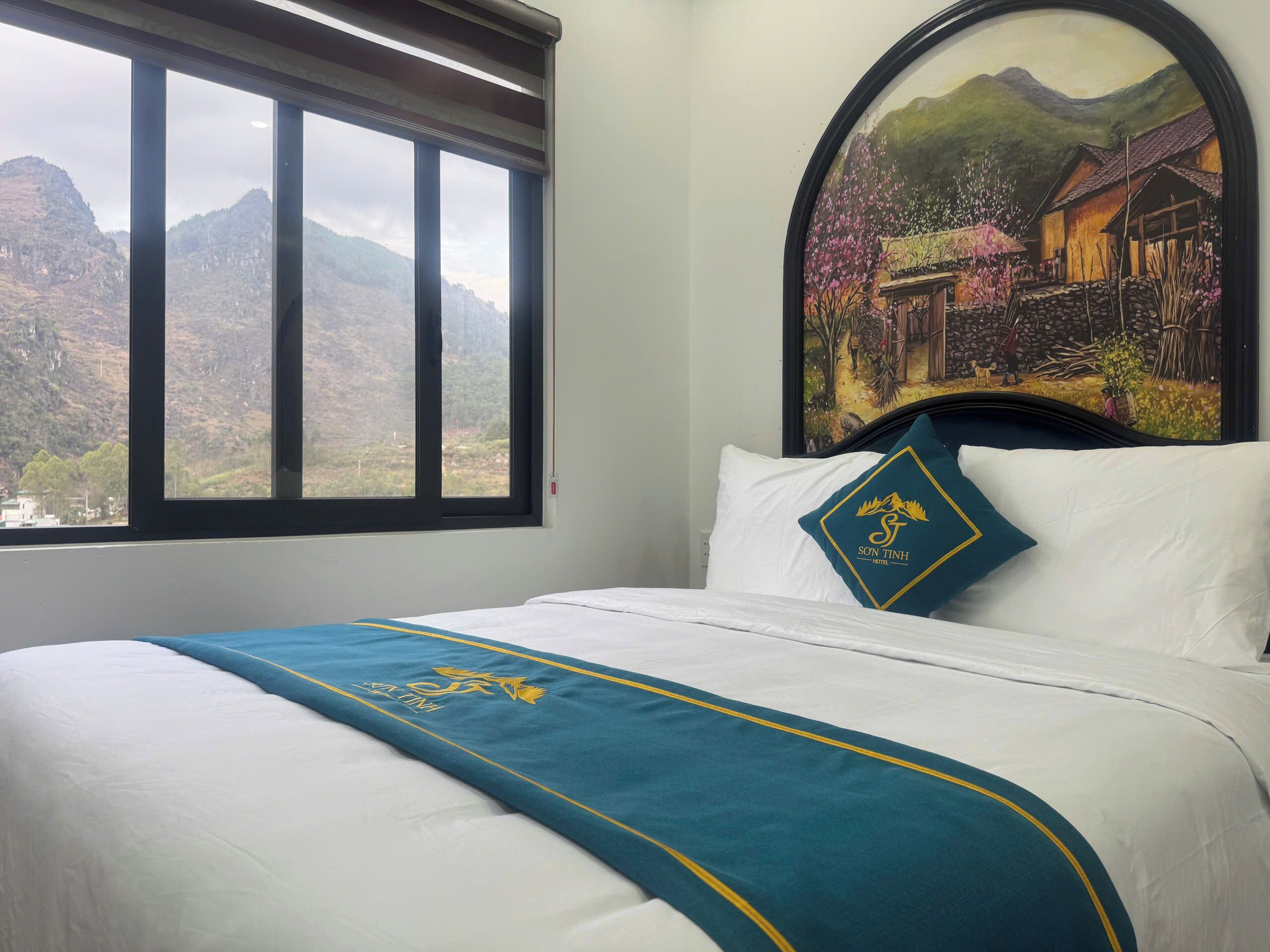 A hotel room with a bed, a window with a mountain view, and a painting of a village.
