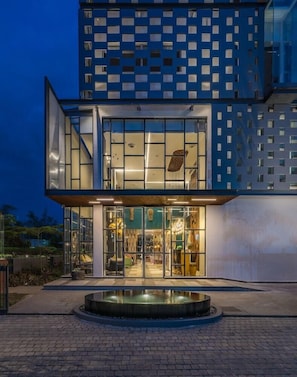 Un bâtiment moderne doté d’une façade en verre, d’une fontaine circulaire à l’avant et d’un mur orné situé à l’étage supérieur.