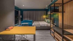 A modern hotel room with a bed, a blue sofa, a dining table, and a wooden shelving unit.