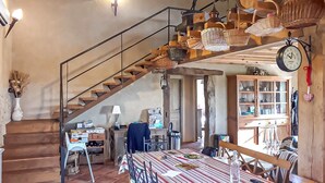 Un interior rústico con una escalera de madera, una zona de comedor con mantel a rayas y un reloj en la pared.