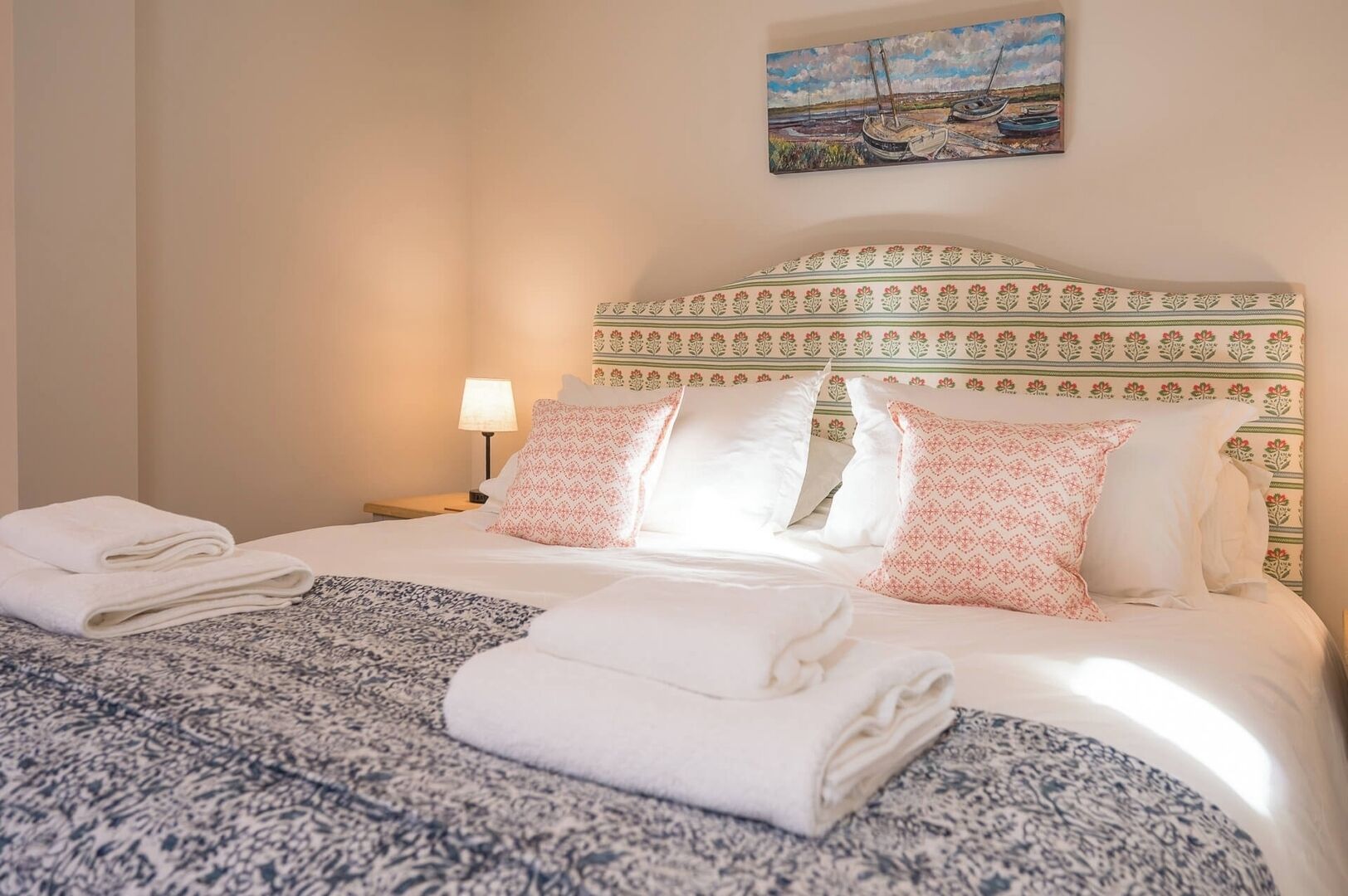 A neatly made bed with white linens and patterned pillows, a bedside lamp, and a painting above the bed.