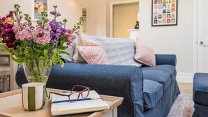 Ein Wohnzimmer mit einer Couch, einem Couchtisch mit Blumenvase, einem Buch und einer Brille.
