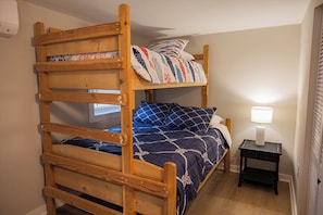 Ein Stockbett mit Holzbettgestell, ein kleiner Nachttisch mit Lampe und ein Fenster mit Jalousien.