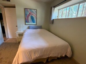 Une chambre avec un lit simple, une table de chevet, une lampe, un tableau abstrait encadré et une fenêtre donnant sur des arbres.