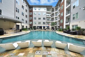 A swimming pool with lounge chairs, a covered area, and a building with multiple floors and balconies.