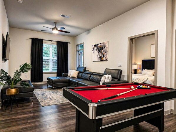 A modern living room with a pool table, a sofa, a ceiling fan, and a large window with curtains.
