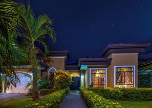 Ein zweigeschossiges Haus mit gut beleuchtetem Eingang, einem Weg zum Garage und einem sternenübersäten Nachthimmel.