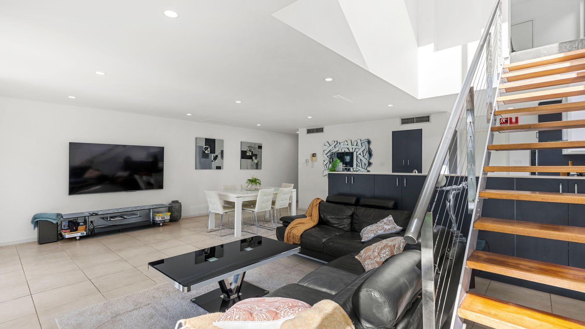 A modern living room with a flat-screen TV, a black leather sofa, a dining area with a white table and chairs, and a staircase with wooden steps.
