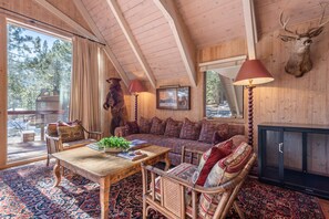 A cozy cabin interior with a large window, a sofa, a wooden table, and a mounted deer head.