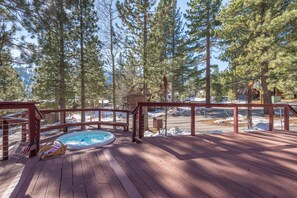 A wooden deck with a hot tub, surrounded by tall pine trees and a view of a snowy landscape.
