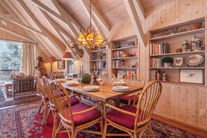 A dining area with a wooden table, red cushioned chairs, a chandelier, and bookshelves with various items.