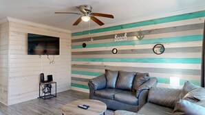 A room with a ceiling fan, a TV mounted on the wall, a sofa with cushions, a small table with a remote control, and a wall with horizontal wooden planks painted in various colors. - Shared Pool, Pet Friendly (Port Aransas)