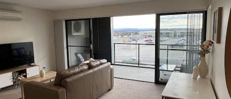 A modern living room with a sofa, a flat-screen TV, a dining table, and a view of a parking lot through large glass doors.