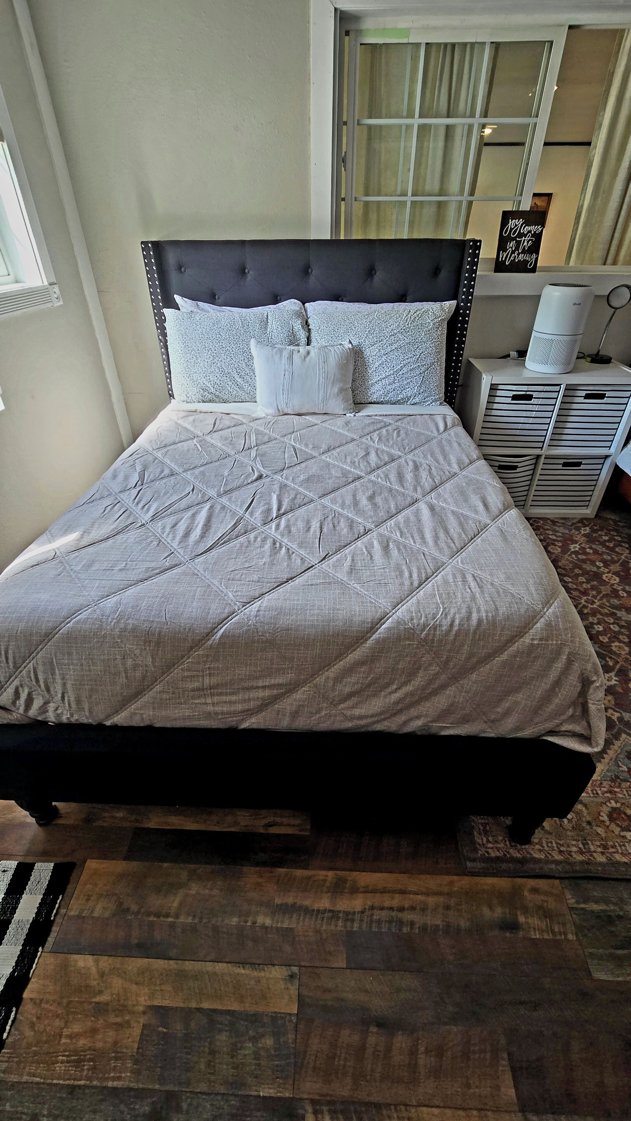 Un lit avec une couvre-lit matelassée grise, une tête de lit sombre et plusieurs oreillers. Il y a une table de chevet avec une lampe et un panneau au-dessus.