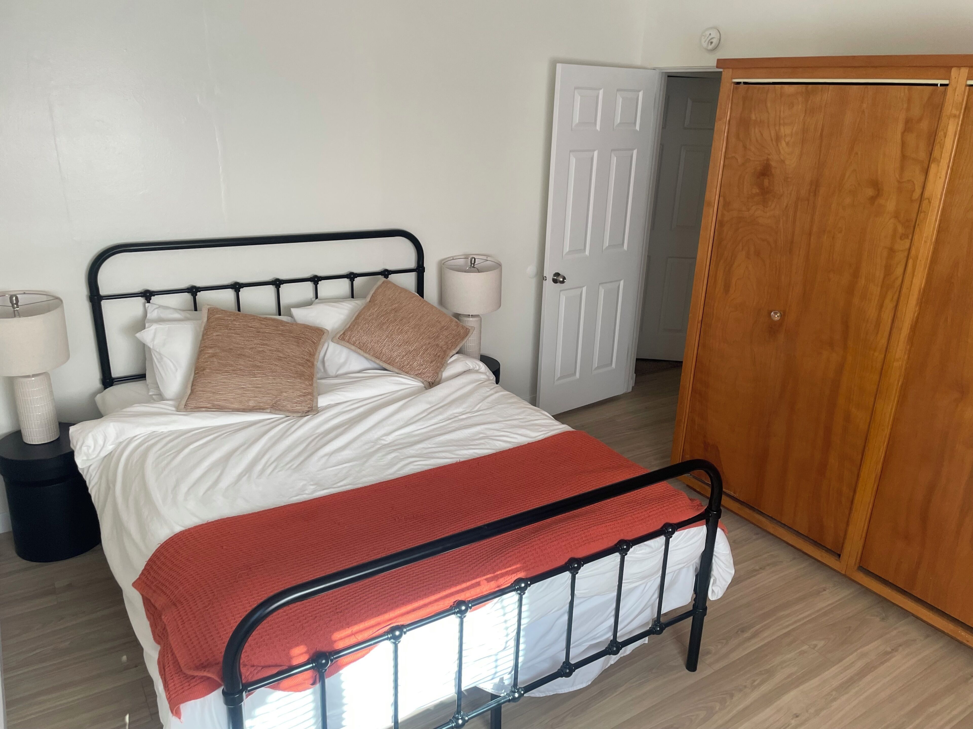 Ein Bett mit Metallgestell, weißer Bettwäsche und zwei Kissen. Im Hintergrund steht ein Holzschrank und eine weiße Tür.