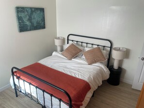 Un letto rifatto con cura, con struttura in metallo, biancheria bianca e una coperta rossa. Sono presenti due lampade da comodino e un cestino nero.