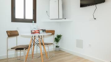 A small room with a white wall, a window, a white air conditioner, a flat-screen TV, a round table with two chairs, and a small potted plant.
