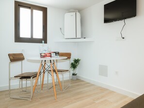 Une petite pièce avec un mur blanc, une fenêtre, un climatiseur blanc, un téléviseur à écran plat, une table ronde avec deux chaises et une petite plante en pot.