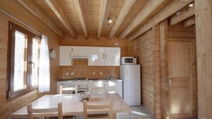 Interior de una cabaña de madera con mesa de comedor, sillas, zona de cocina con armarios y electrodomésticos, y una ventana con cortinas.