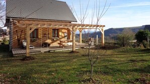 Una casa de madera con un porche cubierto, un árbol pequeño en el patio y montañas al fondo.