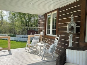 Une terrasse en bois avec des fauteuils à bascule blancs et une table, une décoration en forme de nichoir et une vue sur une cour clôturée.