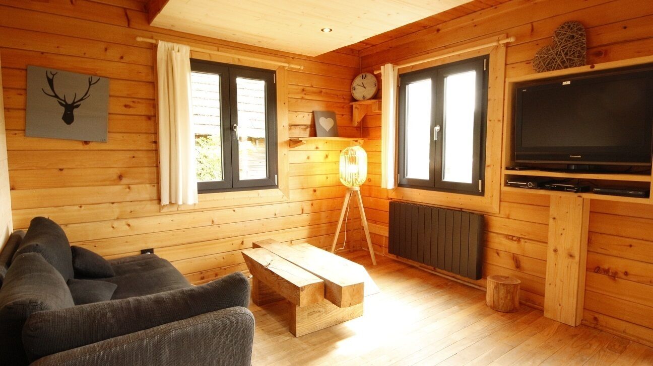Interior de una cabaña de madera con sofá, una mesita de centro de madera, lámpara, televisor y un reloj de pared.