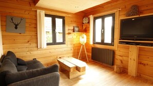A wooden cabin interior with a sofa, a wooden coffee table, a lamp, a television, and a wall clock.