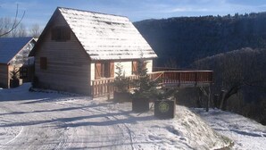 A log cabin with a wooden deck, surrounded by snow-covered ground and a mountainous backdrop.
