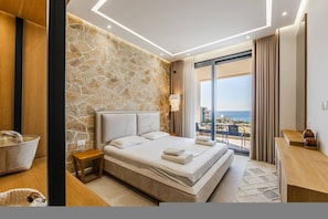 Ein modernes Hotelzimmer mit Steinwand, einem Bett mit weißen Leinen, einem hölzernen Nachttisch, einem Schreibtisch und Blick aufs Meer.