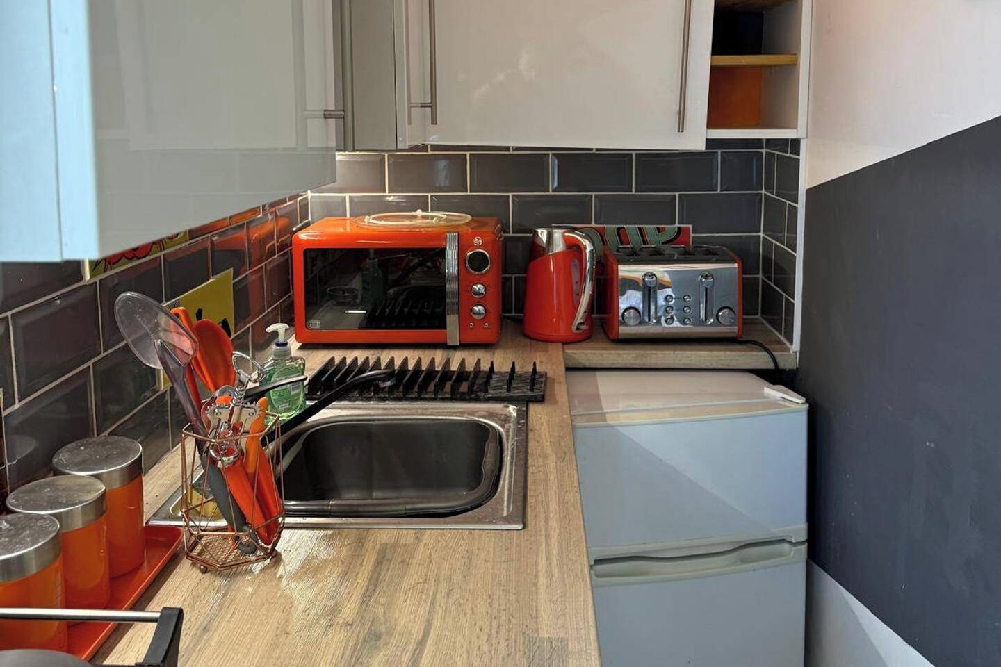 Een kleine keuken met een magnetron, broodrooster en waterkoker, een spoelbak met afdruiprek en kastjes.