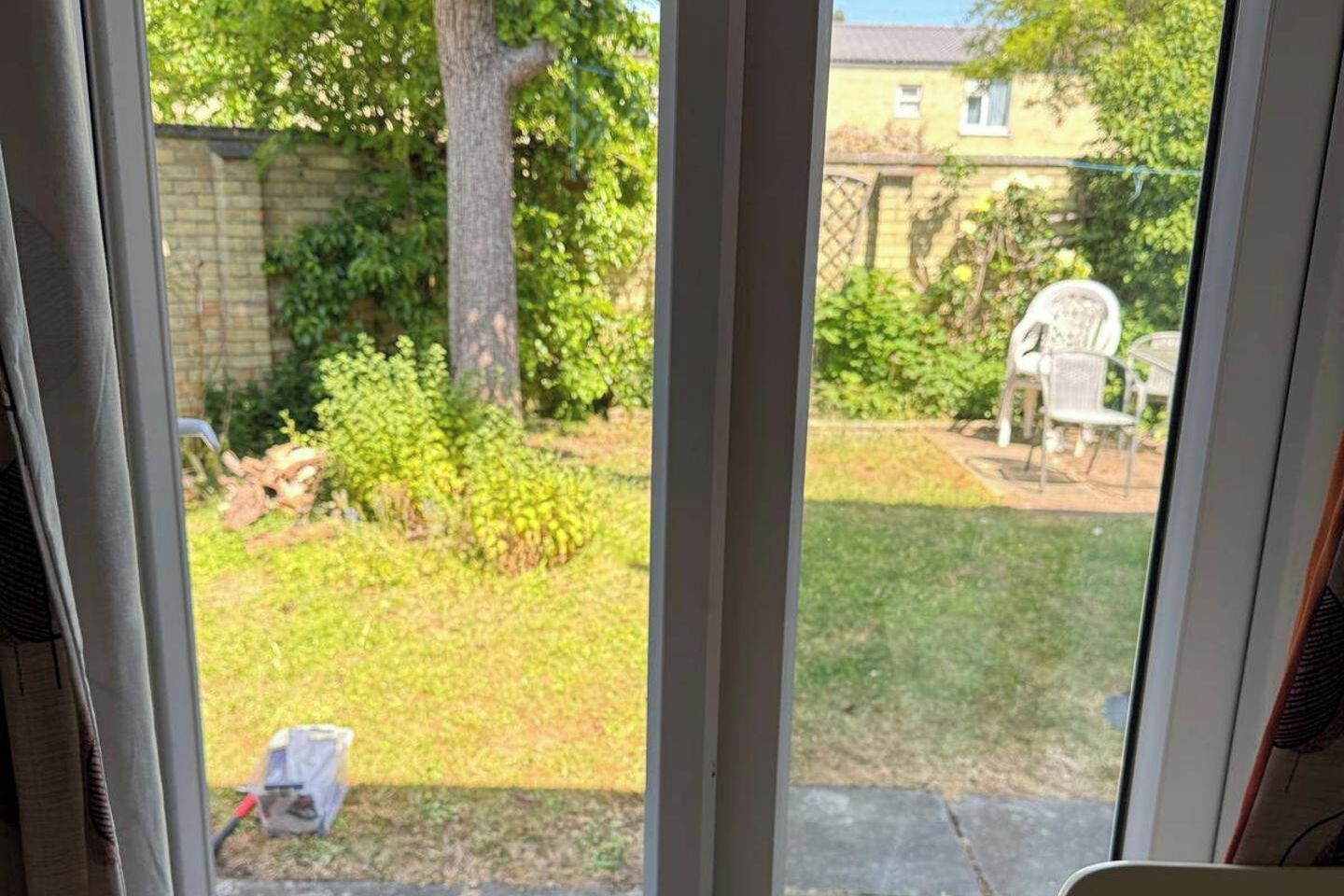 A view through a window of a backyard with a lawn, a tree, and a patio area with chairs.