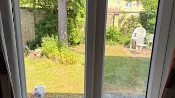 A view through a window of a backyard with a lawn, a tree, and a patio area with chairs.