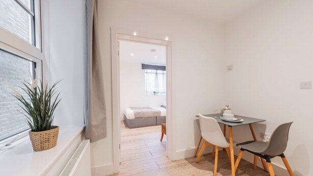 Ein modernes Zimmer mit Essbereich, einem Fenster mit Pflanze und Blick in ein weiteres Zimmer mit Bett.