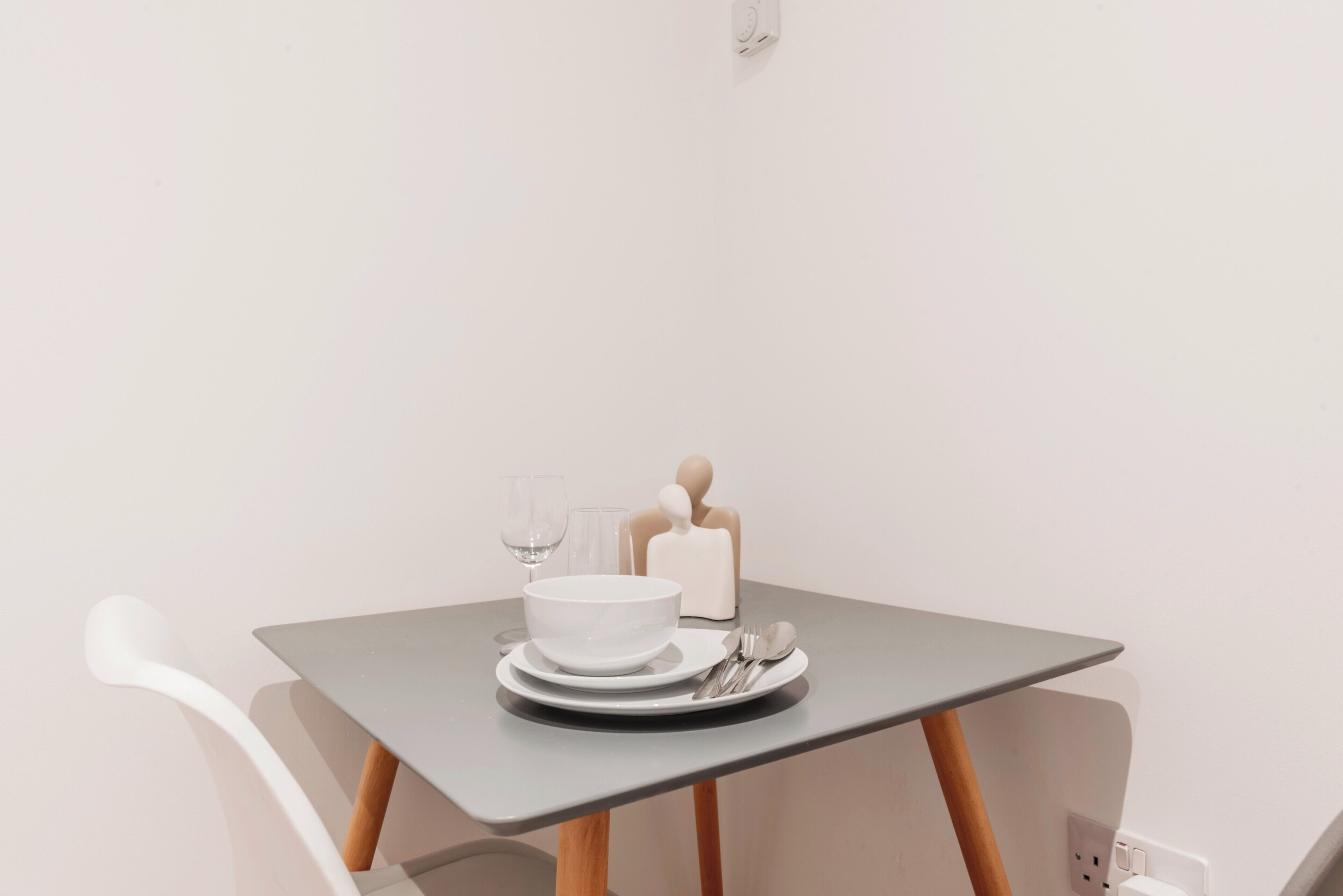 A minimalist dining table with a bowl, plates, cutlery, and glasses, set against a plain wall.
