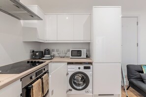A compact kitchen with a microwave, toaster, and washing machine, a small oven, and a black sofa in the living area.