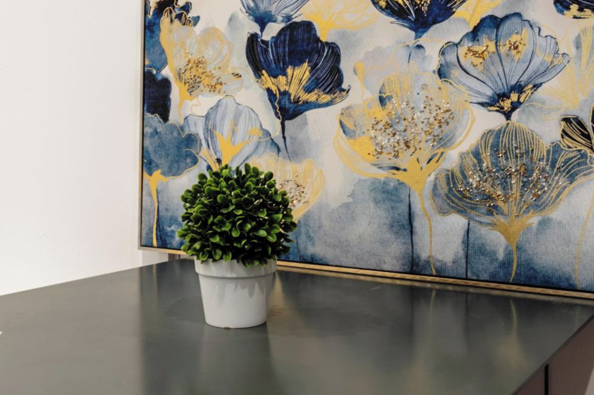 Una obra de arte floral en un marco colgada en una pared, una planta en maceta sobre una mesa y una encimera de color oscuro.