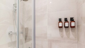 Une douche fermée par des parois en verre, avec un pommeau de douche mobile, trois bouteilles de shampoing fixées au mur et un sol carrelé.