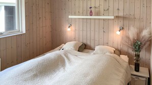 Ein Schlafzimmer mit einer holzverkleideten Wand, einem weißen Bett mit floralem Überwurf, zwei Nachttischlampen und einer kleinen Ablage mit einer Vase.