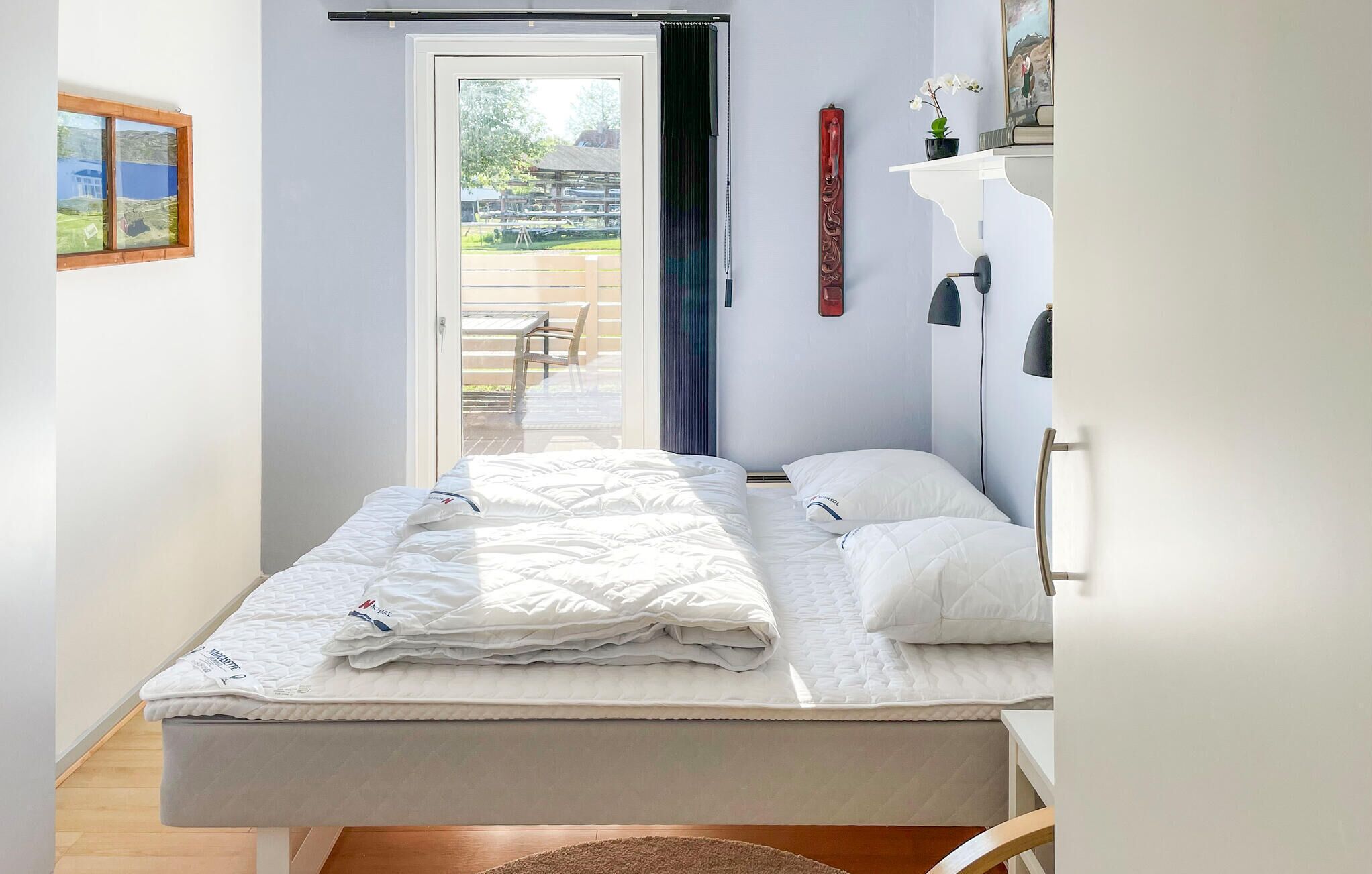 Une chambre avec un lit, une porte donnant sur un espace extérieur, une petite table et une étagère fixée au mur, sur laquelle se trouve une plante.