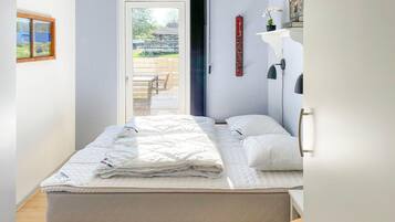 Une chambre avec un lit, une porte donnant sur un espace extérieur, une petite table et une étagère fixée au mur, sur laquelle se trouve une plante.
