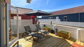 Une terrasse en bois avec deux chaises longues et une table, un bac à fleurs en bois contenant des plantes et une clôture blanche.