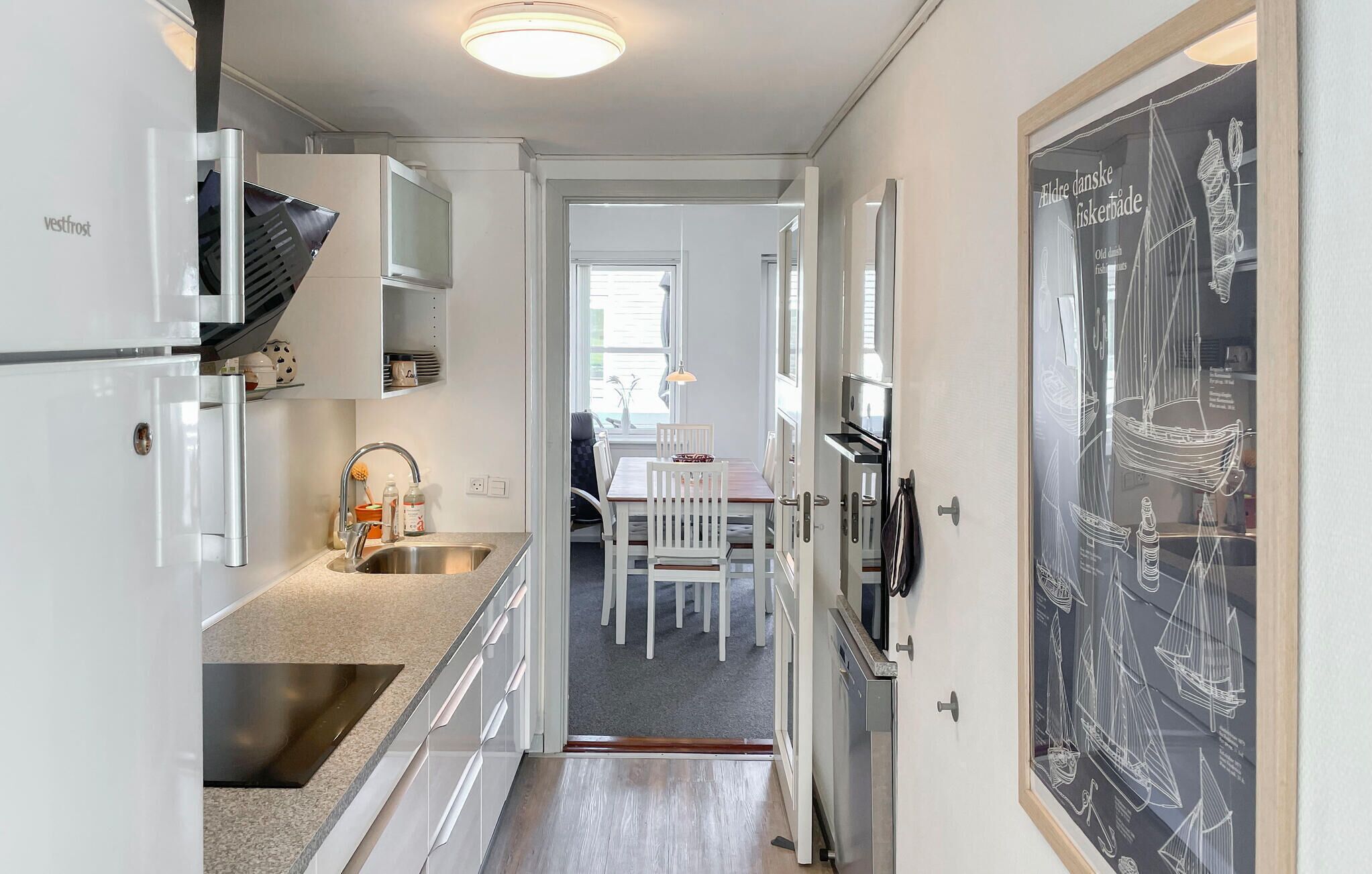 Un couloir étroit, avec une cuisine d’un côté, un coin repas avec une table et des chaises, et une grande image encadrée accrochée au mur.