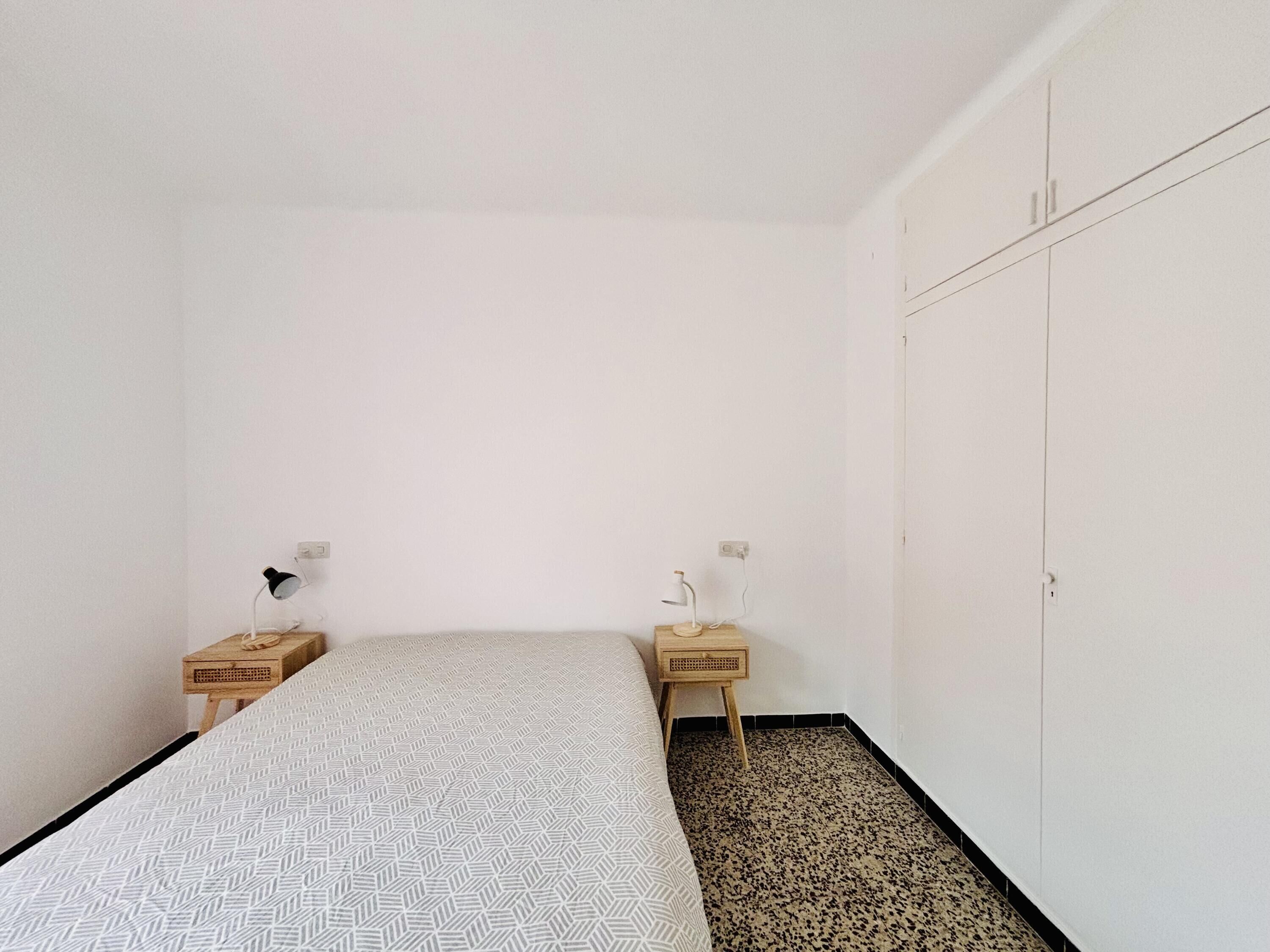 A small, minimalist bedroom with a bed, two nightstands, a wardrobe, and a patterned floor.