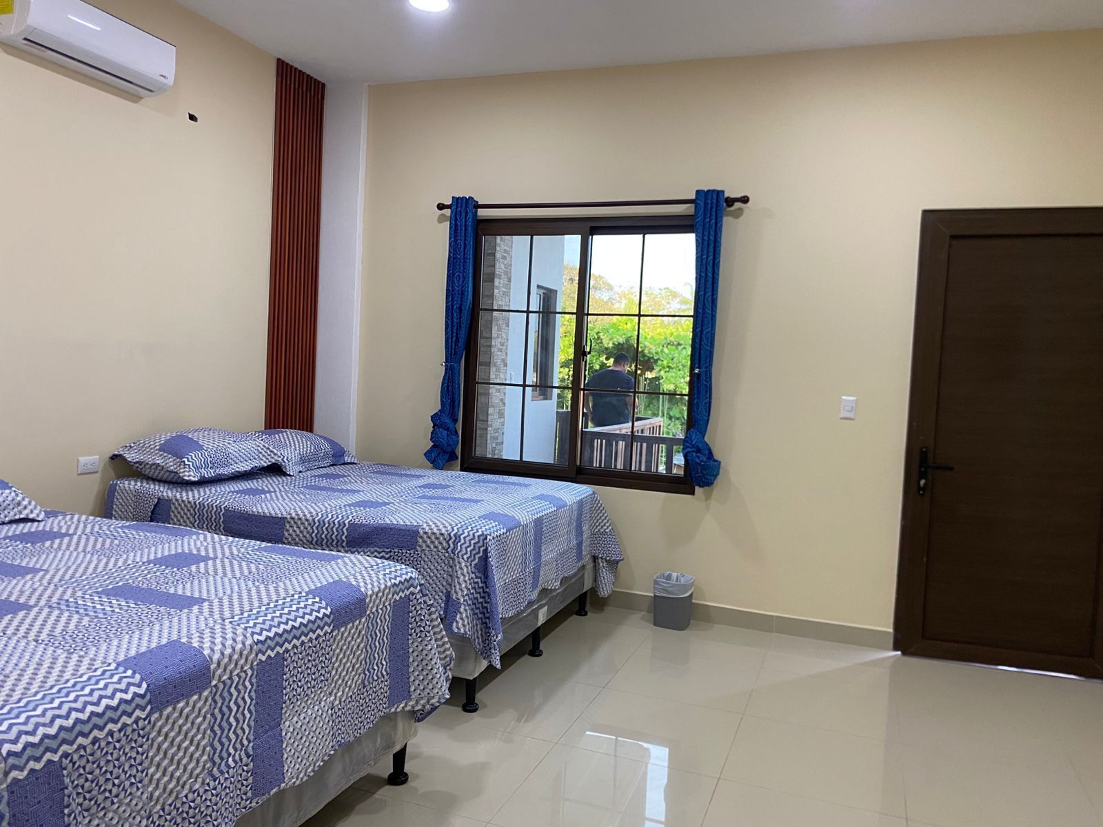 A room with two beds, a window with curtains, a door, and an air conditioning unit.