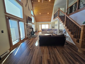 A spacious living area with a stone fireplace, wooden flooring, a staircase with a wooden railing, and large windows allowing natural light.