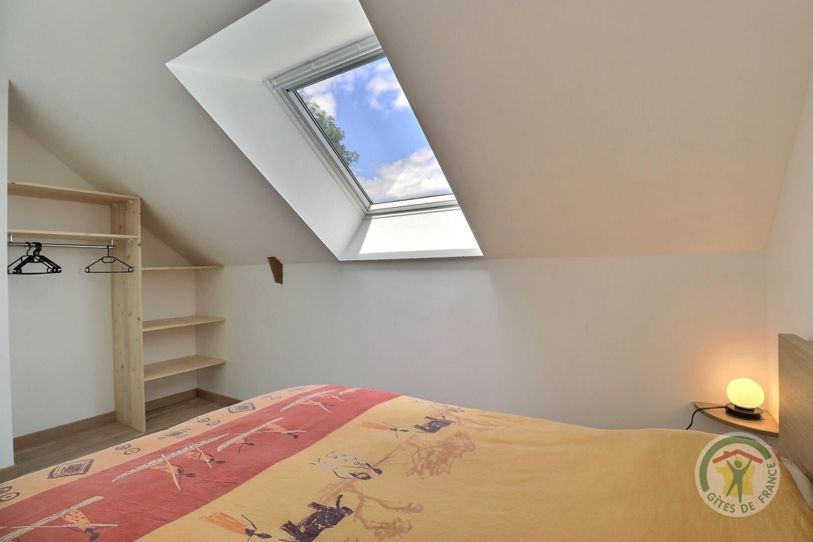 A bedroom with a skylight, a bed with a patterned bedspread, a wooden wardrobe, and a nightstand with a lamp.