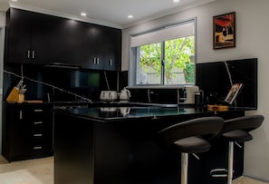 A modern kitchen with black cabinets, a marble countertop, and a large window. - Koblenz Haus - “Heart of the Barossa” - (New Mgmt) (Tanunda)