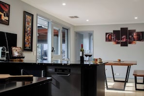 A modern kitchen with a built-in dishwasher, a dining area with a wooden table, and artwork on the walls. - Koblenz Haus - “Heart of the Barossa” - (New Mgmt) (Tanunda)