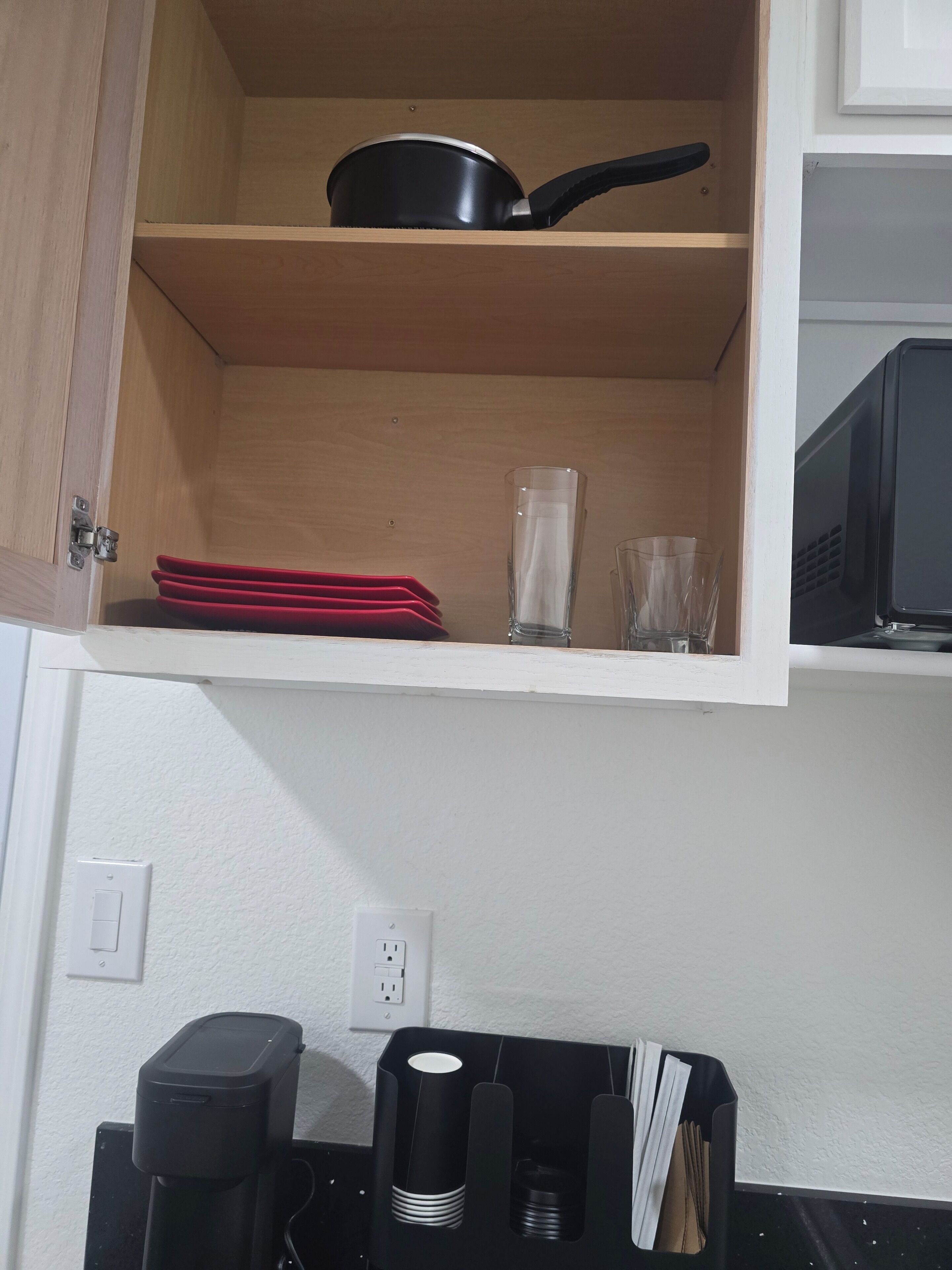 A kitchen with a microwave, a coffee machine, and a shelf with a frying pan and red plates.