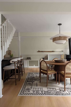 A dining area with a wooden table, chairs, a woven rug, and a hanging light fixture.